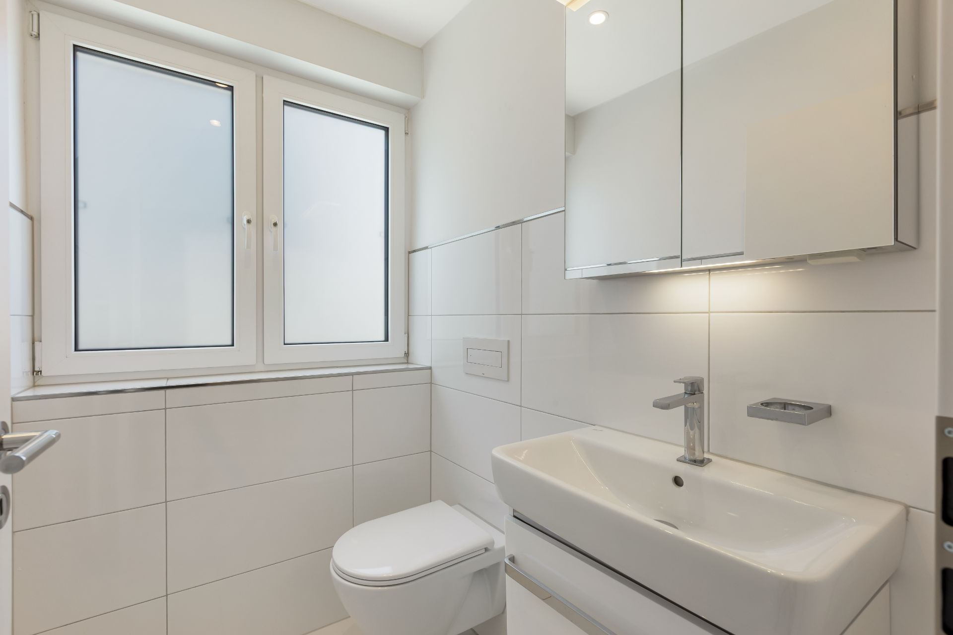 Helles Badezimmer mit großem Fenster, weißer Toilette und Waschbecken mit Spiegel.