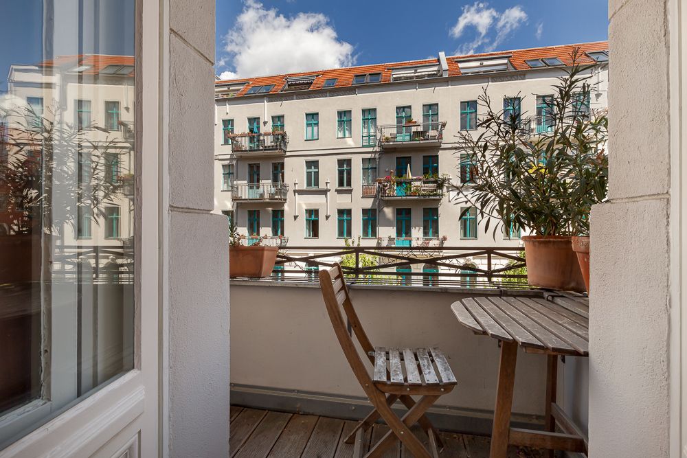 Balkon mit Holztisch, zwei Stühlen und Pflanzen, Blick auf ein Wohnhaus mit Balkonen.