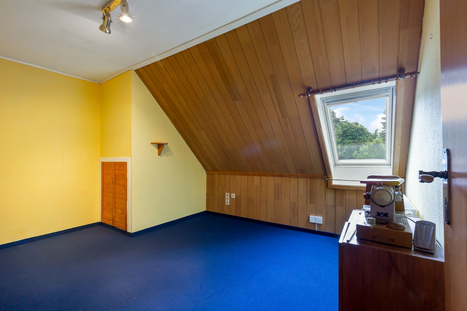 Dachzimmer mit gelben Wänden, schräger Holzdecke, blauem Teppich und Fenster mit Ausblick.