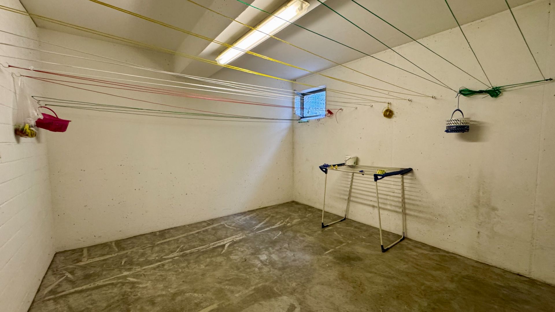 Kellerraum mit Wäscheleinen an der Decke, einem kleinen Fenster und einem Wäscheständer.