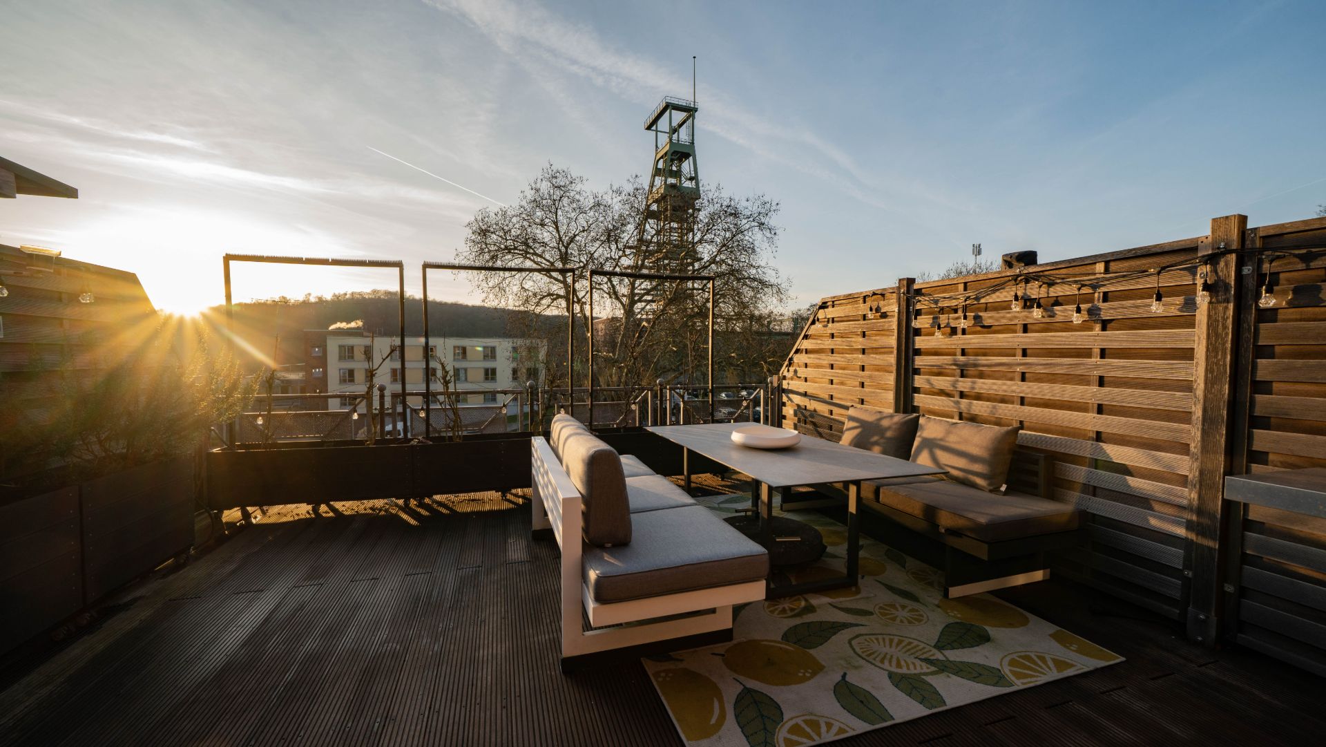 Moderne Dachterrasse mit Sitzbank, Tisch, Teppich und Holzwand bei Sonnenuntergang.