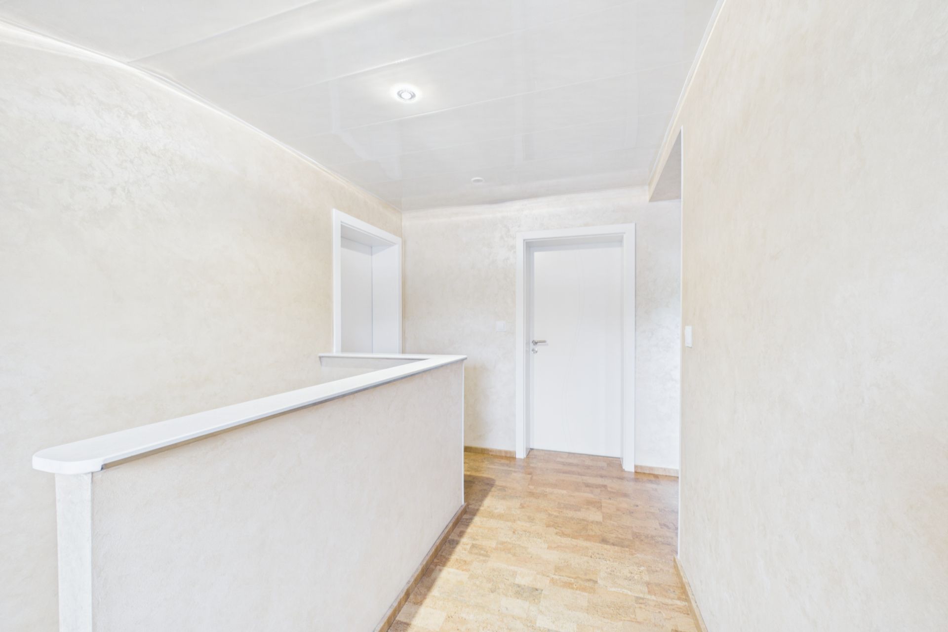 Heller Flur mit weißer Tür, beigem Boden und Wand, Deckenlampe und Geländer.