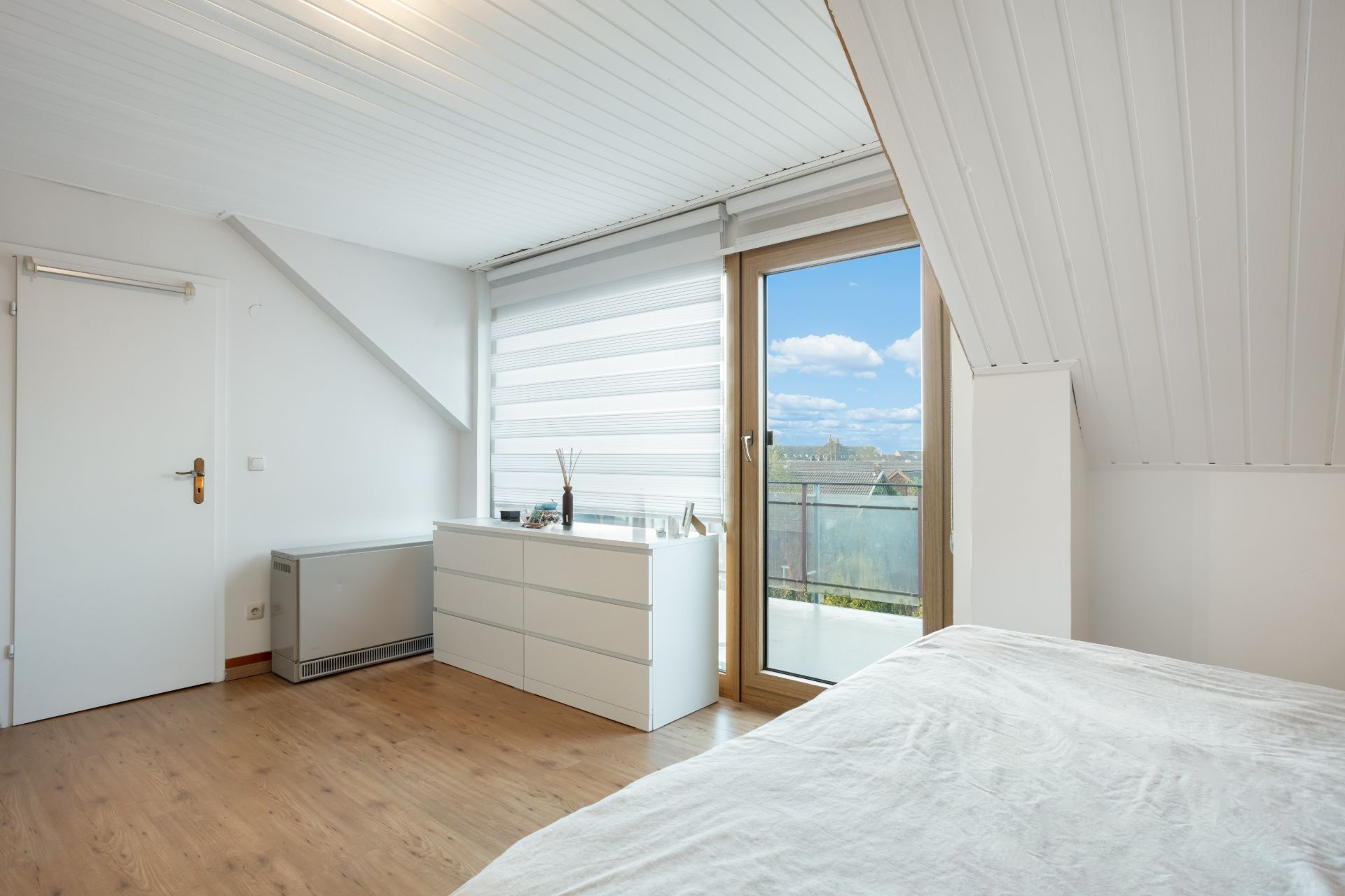 Helles Schlafzimmer mit Holzfußboden, Kommode, Tür und großem Fenster mit Blick nach draußen.