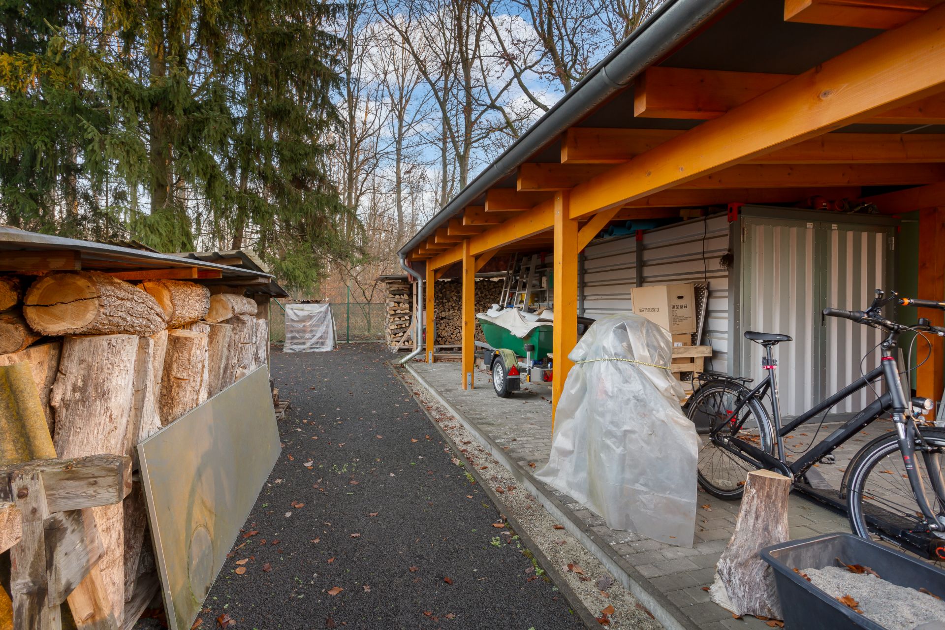 Überdachter Fahrrad- und Geräteschuppen neben einem Holzlager im Garten.
