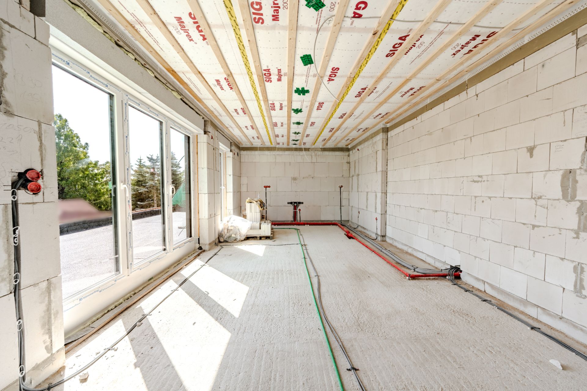 Heller Raum im Rohbau mit großen Fenstern und sichtbaren Leitungen auf Boden und Decke.