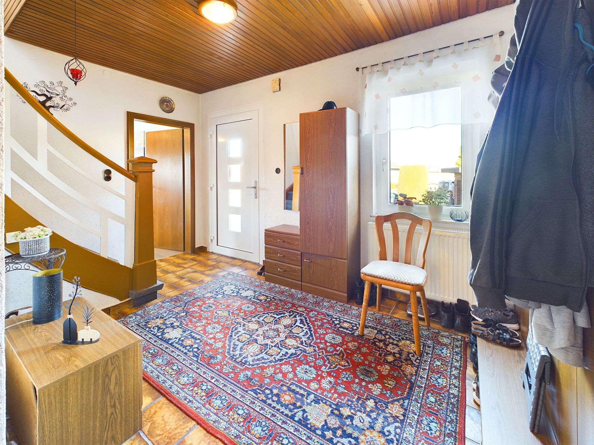Flur mit Treppe, Teppich, Garderobe, Stuhl und Fenster mit Gardine.