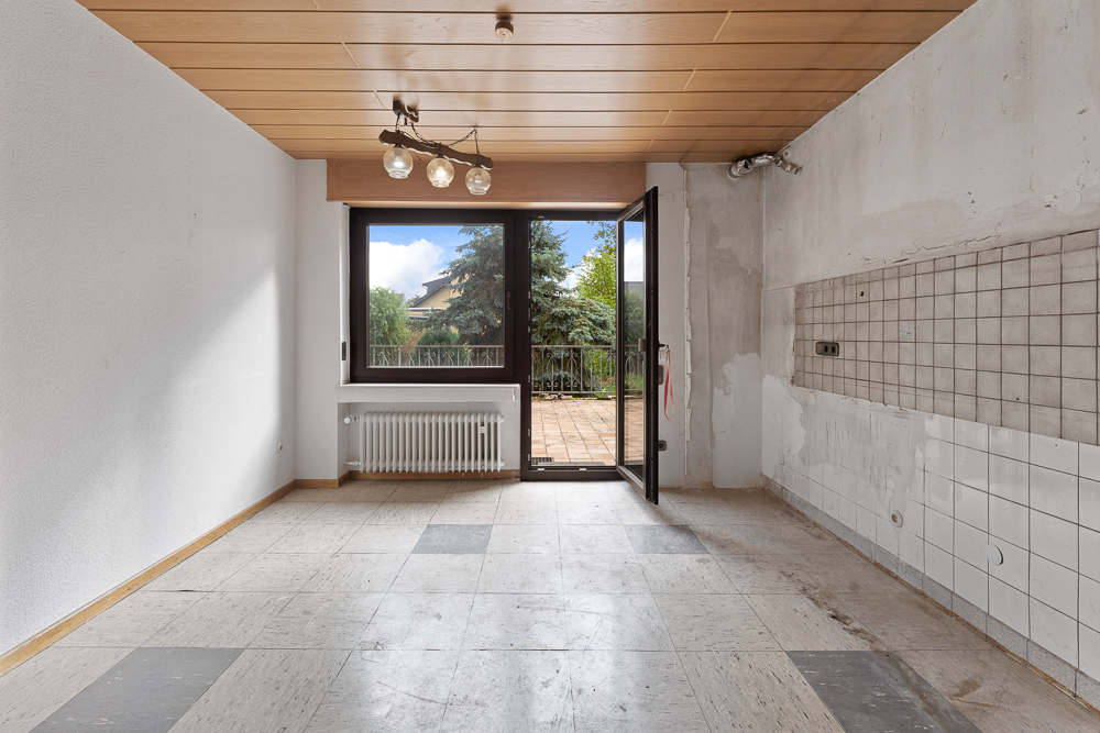 Leerer Raum mit Fliesenboden, großem Fenster und Tür zur Terrasse, teils geflieste Wand.