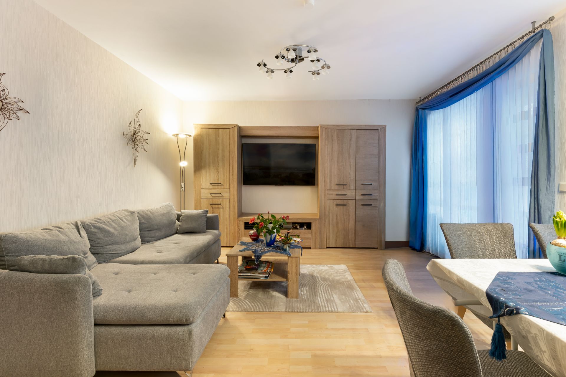 Helles Wohnzimmer mit grauem Sofa, Couchtisch, TV-Schrank und Esstisch am Fenster mit blauen Vorhängen.