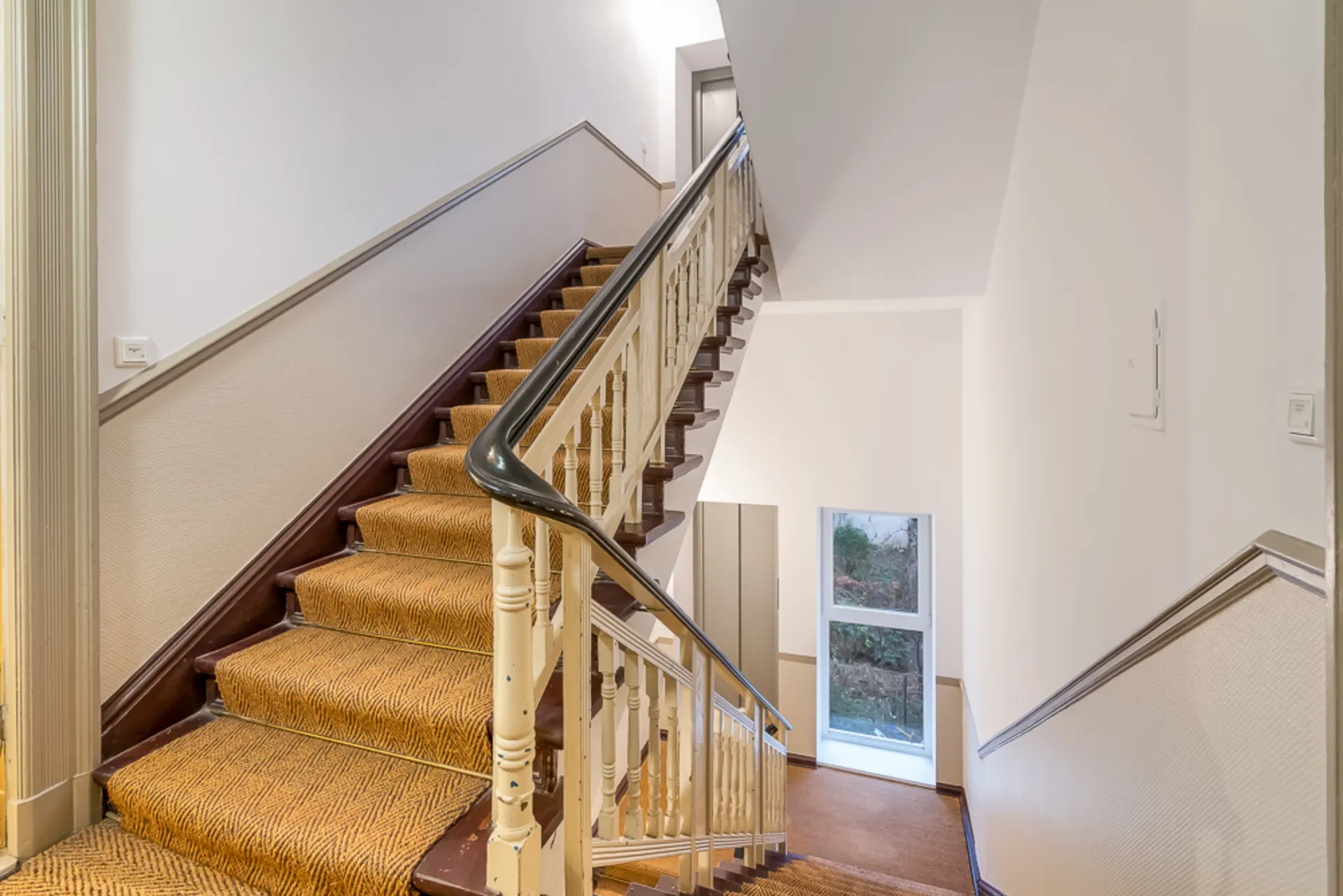 Helles Treppenhaus mit Teppichtreppe, weißen Wänden und Fenster im Erdgeschoss.