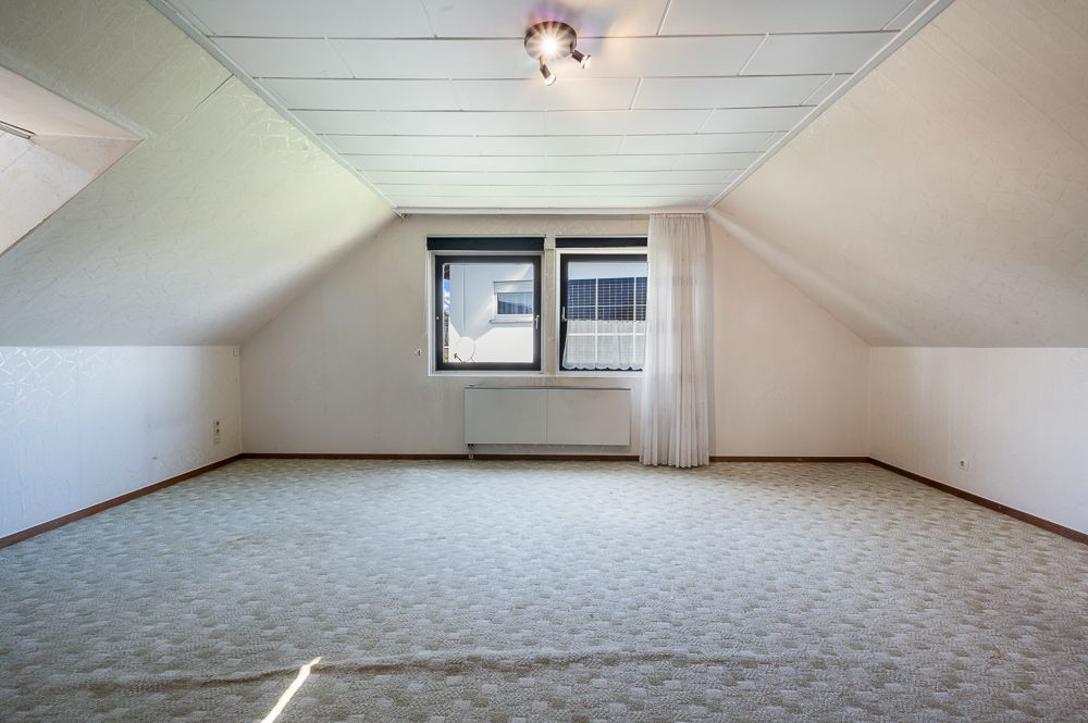 Heller leerer Dachbodenraum mit zwei Fenstern, Teppichboden und schrägen Wänden.