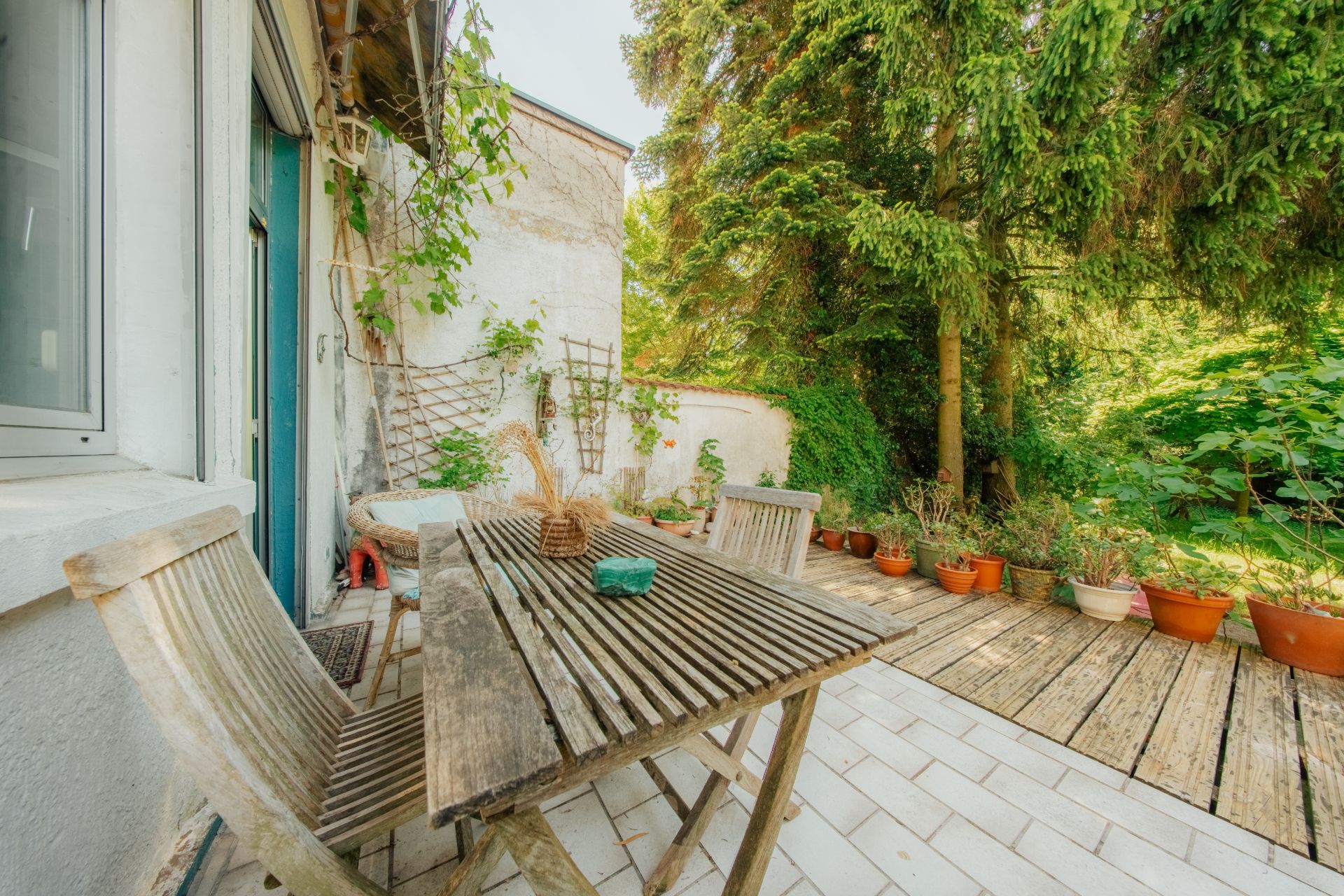 Holztisch mit Stühlen auf Terrasse, viele Pflanzen und Bäume im Garten.