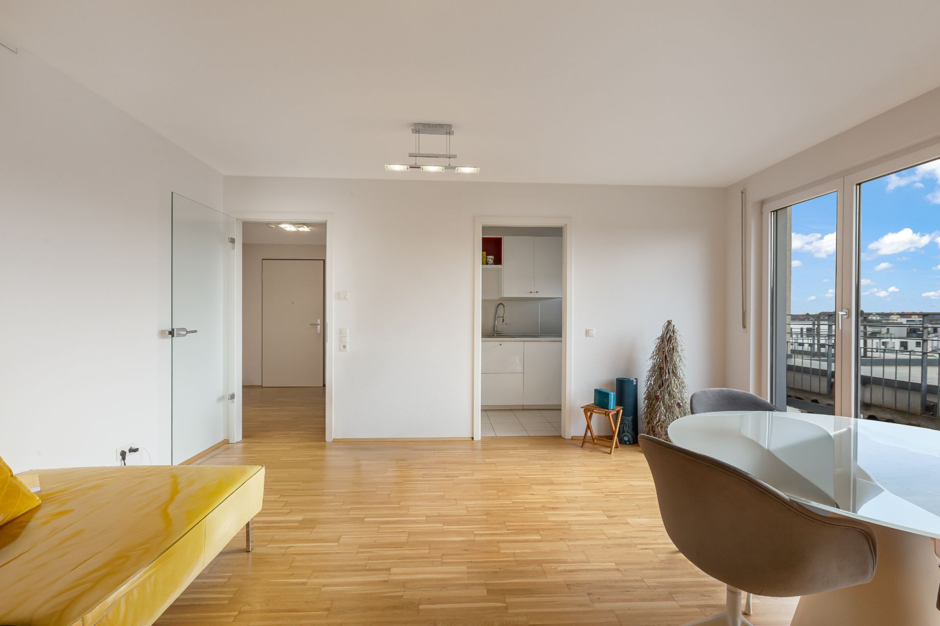 Helles Wohnzimmer mit Glastür, Esstisch, gelber Bank und Zugang zur kleinen Küche.