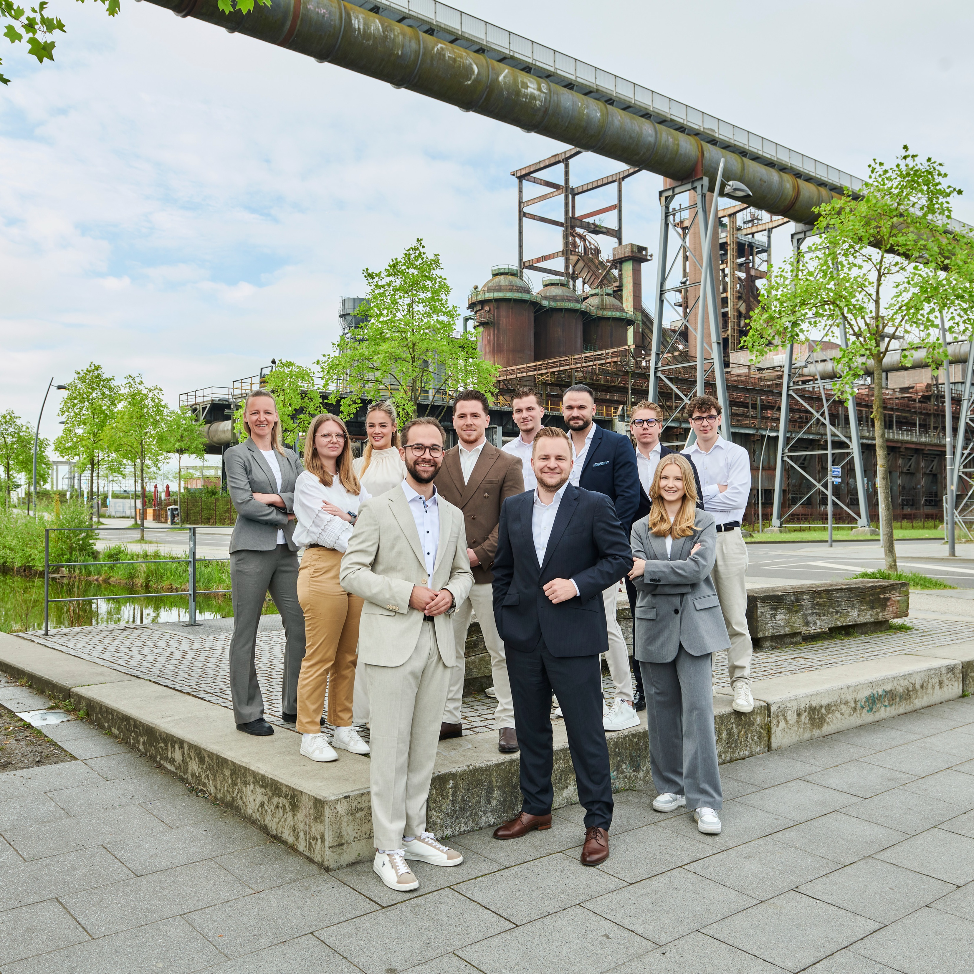 Eine Gruppe von Menschen in Anzügen steht draußen vor einer alten Industrieanlage.