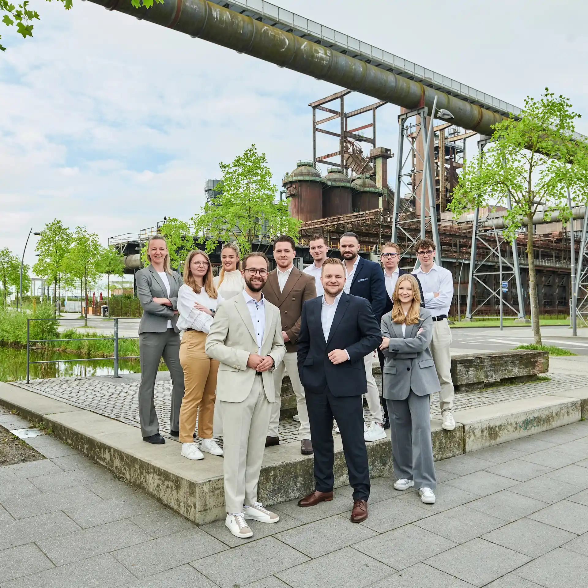 Eine Gruppe von Menschen in Anzügen steht draußen vor einer alten Industrieanlage.