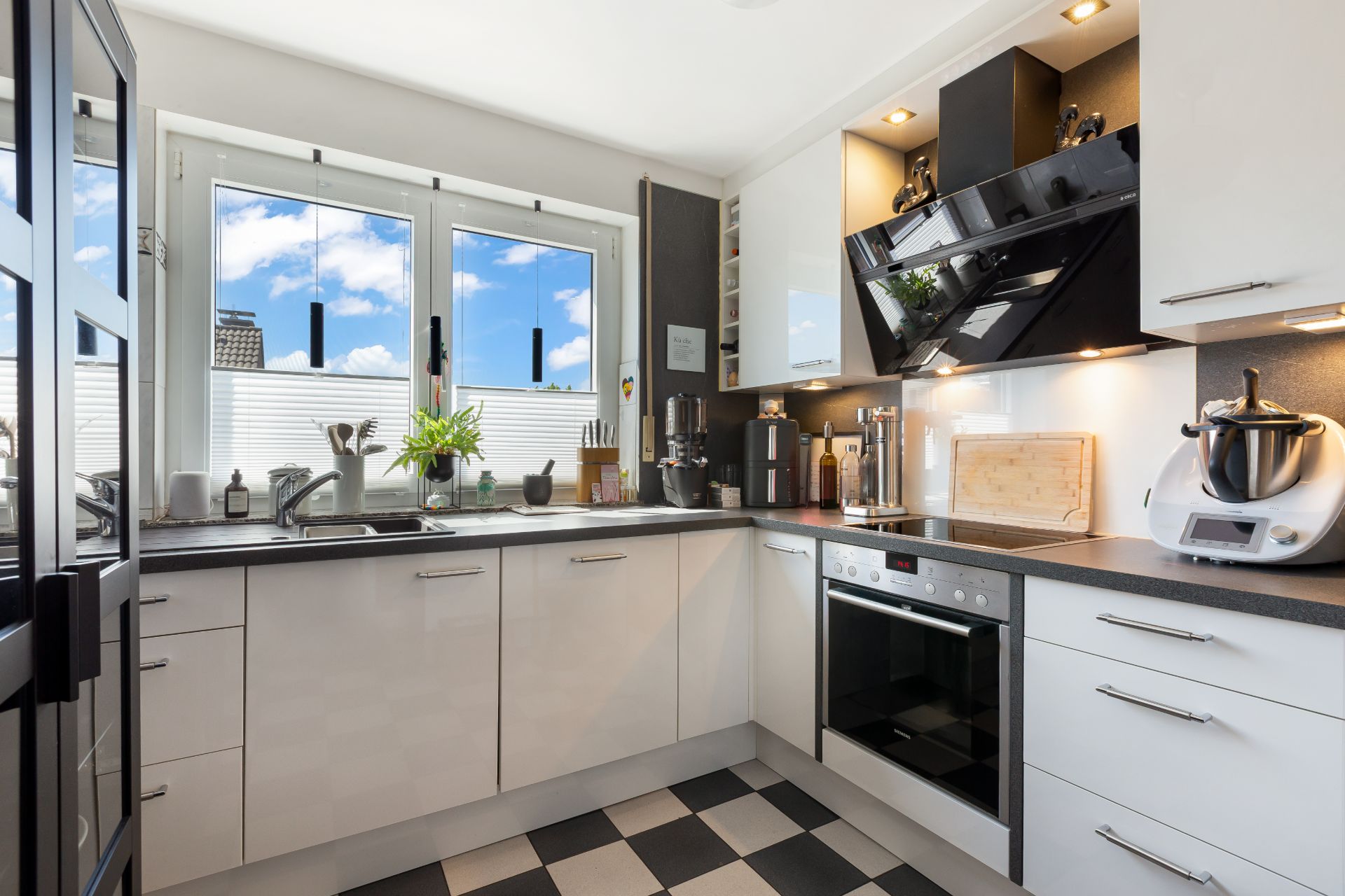 Moderne Küche mit weißen Schränken, schwarzen Arbeitsflächen und kariertem Boden, großes Fenster mit Blick nach draußen.