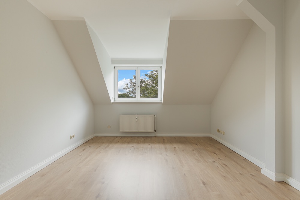 Leerer Raum mit Dachschräge, hellem Holzboden und Fenster mit Blick ins Grüne.