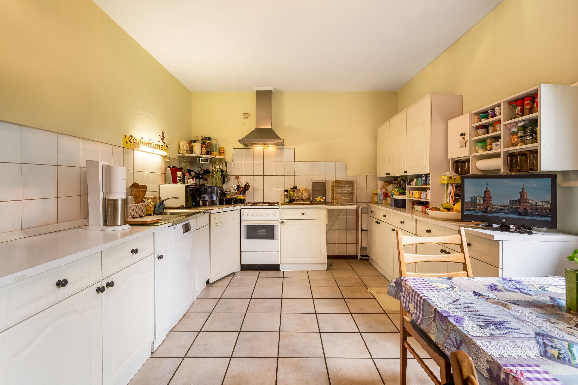 Geräumige Küche mit weißen Schränken, Esstisch, Herd, Dunstabzugshaube und Fernseher.