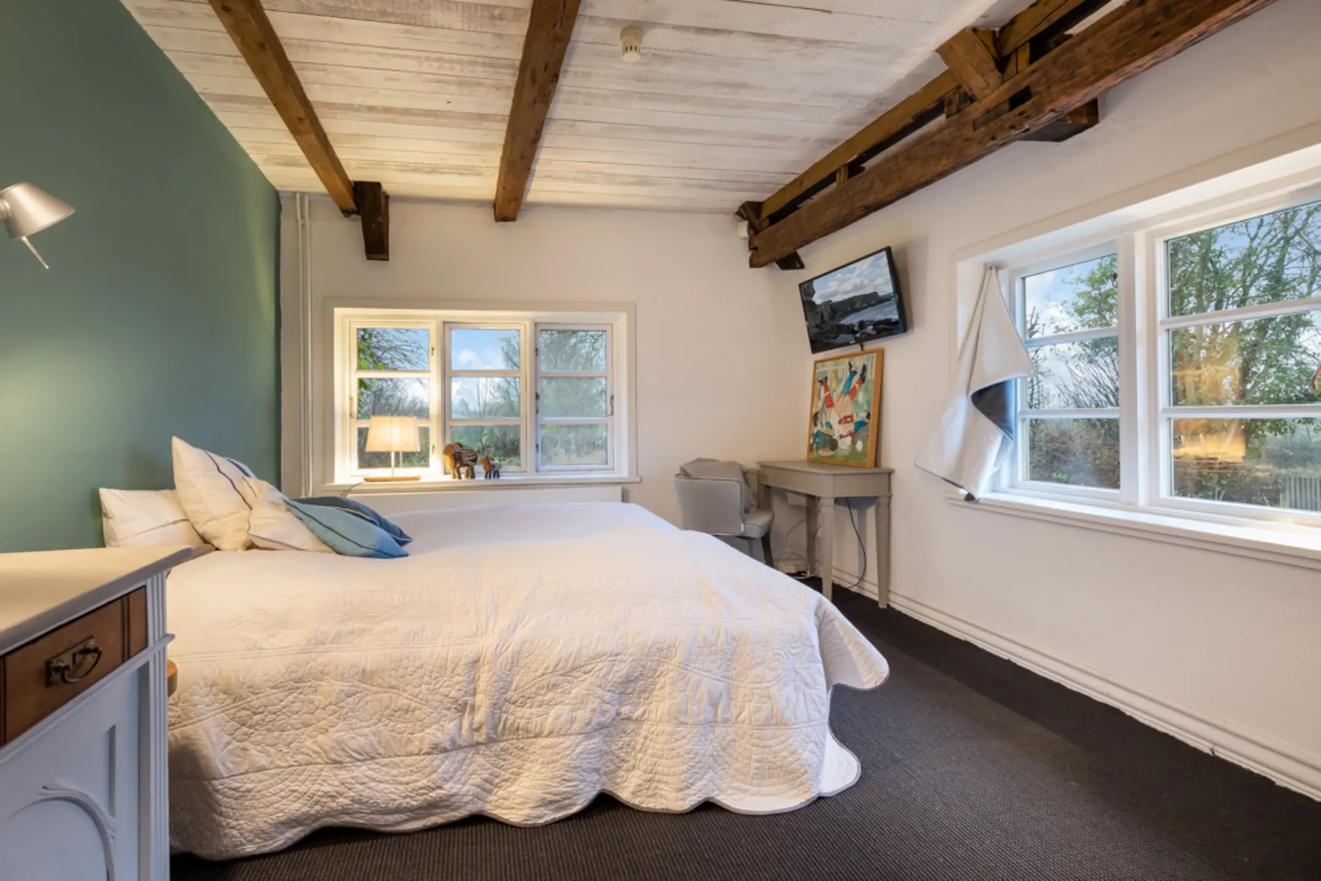 Gemütliches Schlafzimmer mit Doppelbett, Holzbalkendecke, großen Fenstern und Schreibtisch.