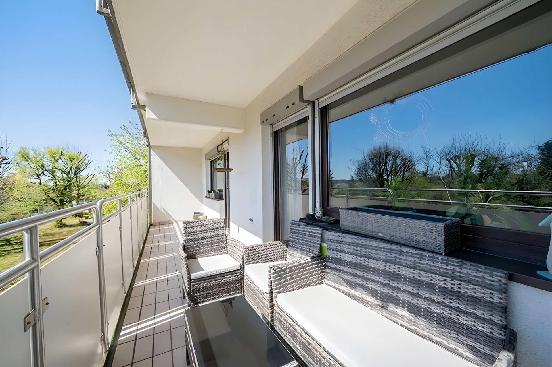 Langer Balkon mit Sitzmöbeln, Glastisch und Blick auf Bäume im Garten.