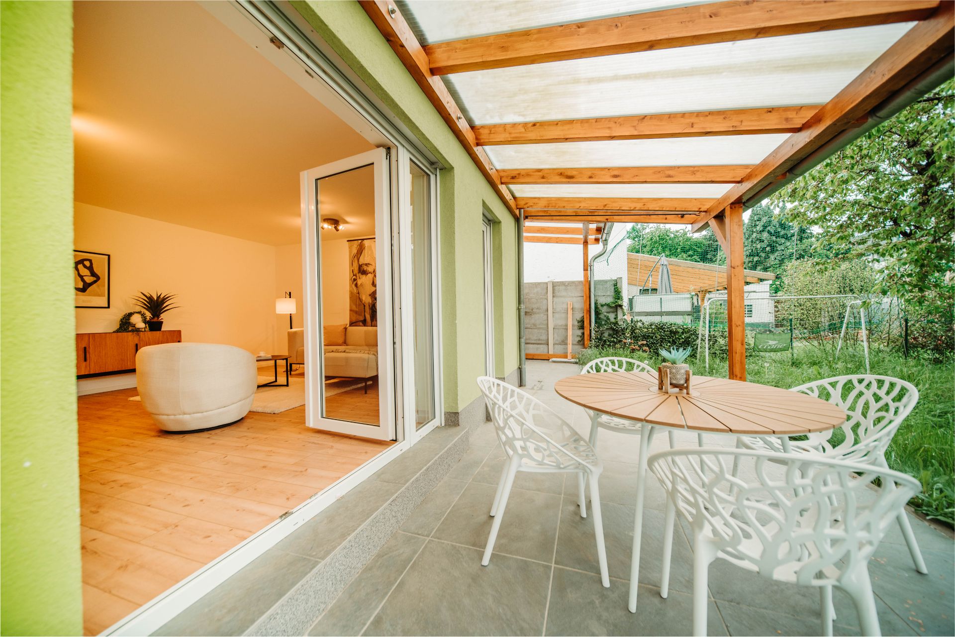 Überdachte Terrasse mit Holztisch und weißen Stühlen, Zugang zum Wohnzimmer mit moderner Einrichtung.