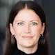 Frau mit langen braunen Haaren steht in schwarzem Outfit mit verschränkten Armen vor Glastür.