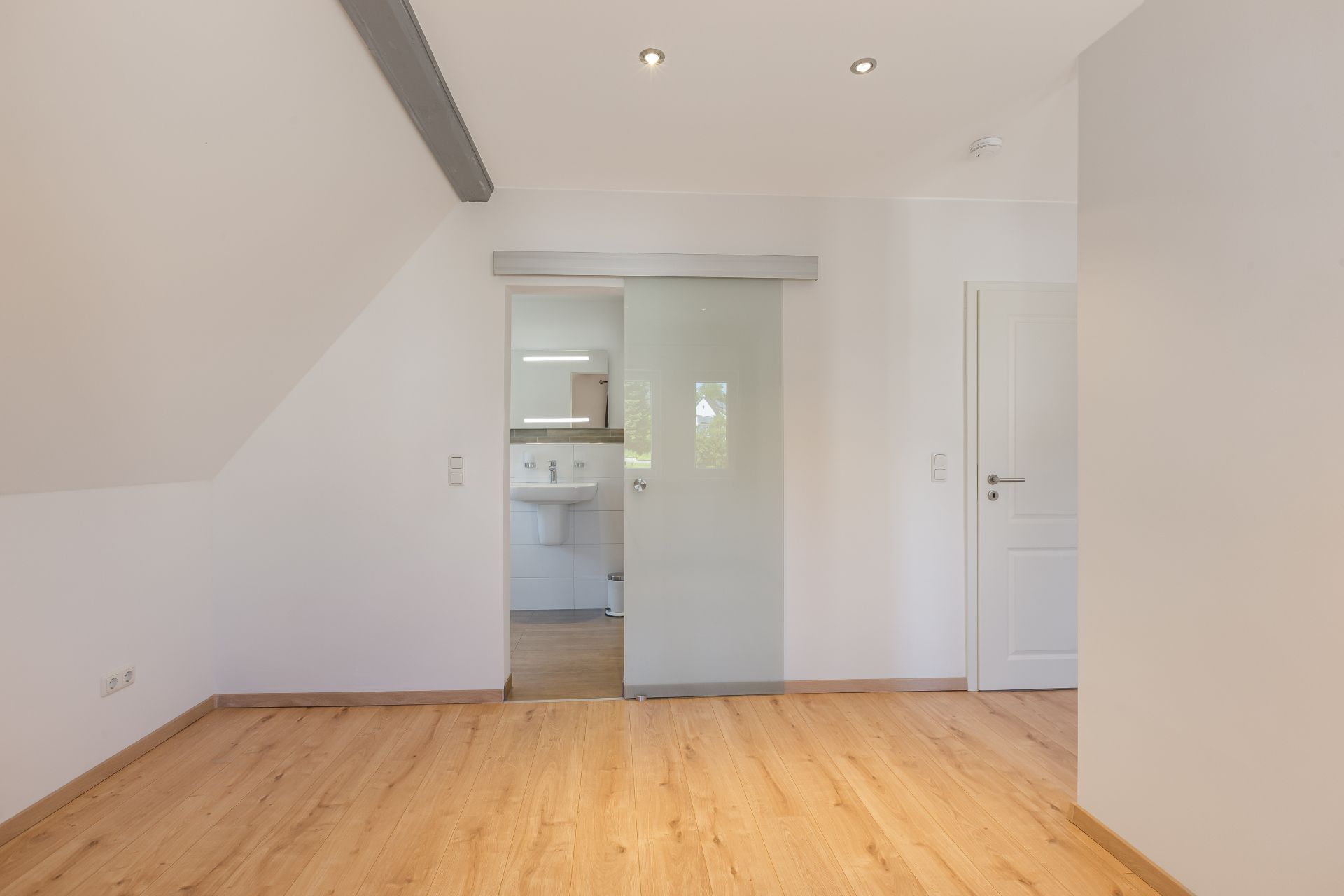 Heller Raum mit Holzfußboden, Schiebetür aus Glas zum Badezimmer, weiße Wände und Decke.