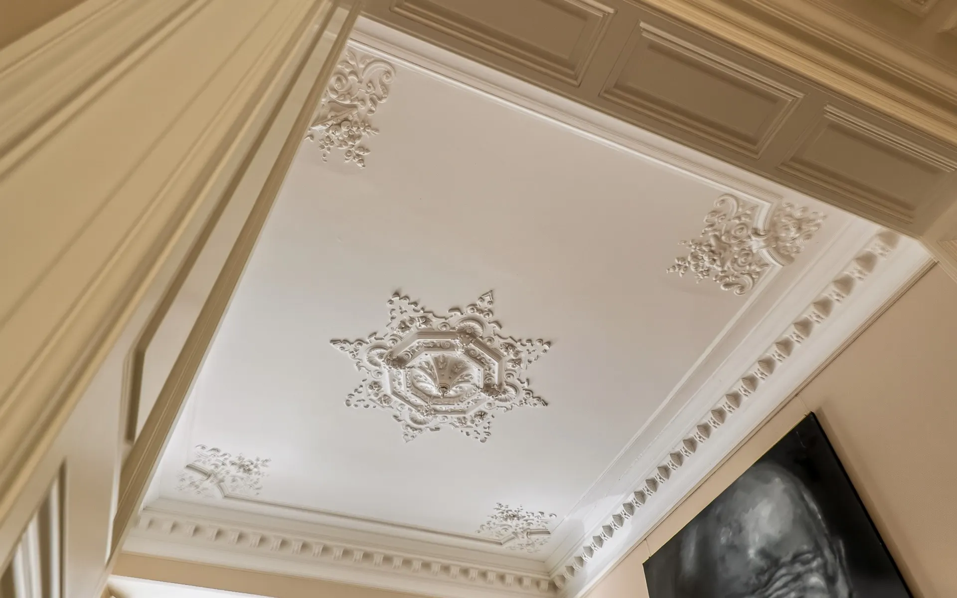 Weiße Stuckdecke mit dekorativen Verzierungen und einem Gemälde an der Wand sichtbar.