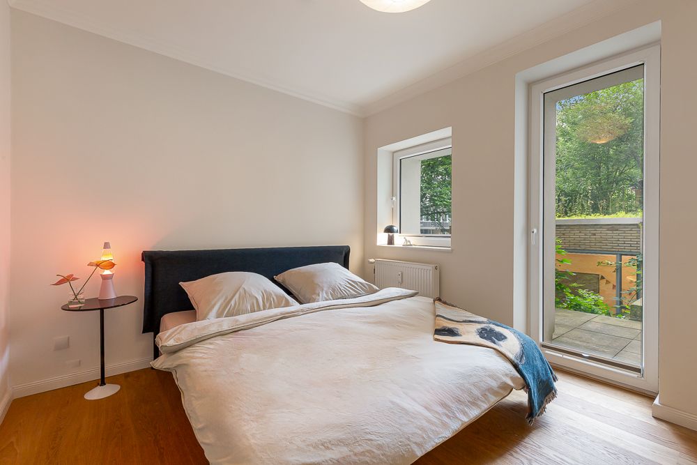 Schlafzimmer mit Doppelbett, Nachttischlampe, Fenster und Tür mit Blick nach draußen.