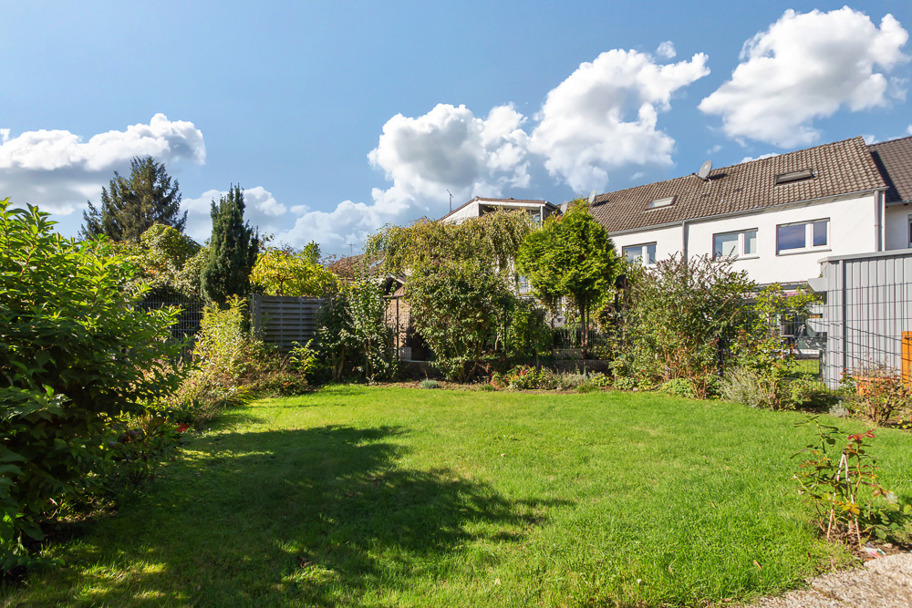 Gepflegter Garten mit Rasenfläche und Büschen, im Hintergrund Reihenhäuser unter blauem Himmel.