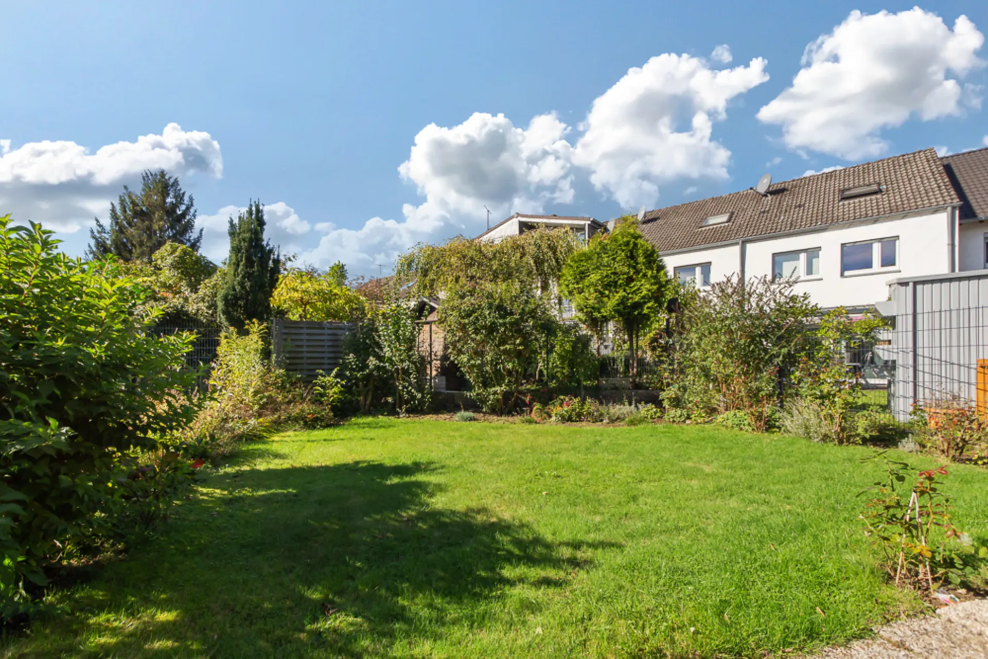 Gepflegter Garten mit Rasenfläche und Büschen, im Hintergrund Reihenhäuser unter blauem Himmel.
