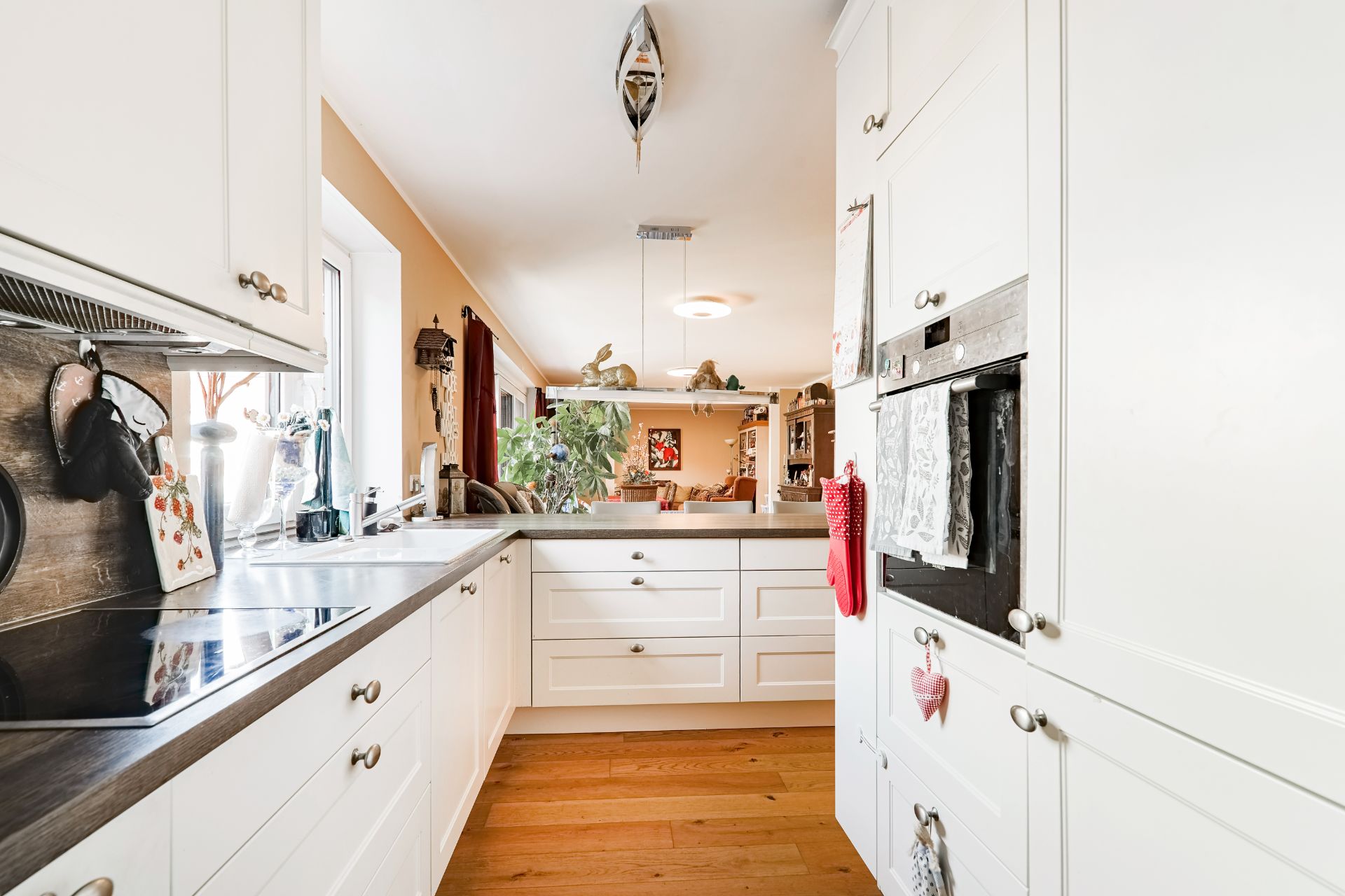 Helle Küche mit weißen Schränken, Holzfußboden und Geräten, Blick ins Wohnzimmer.