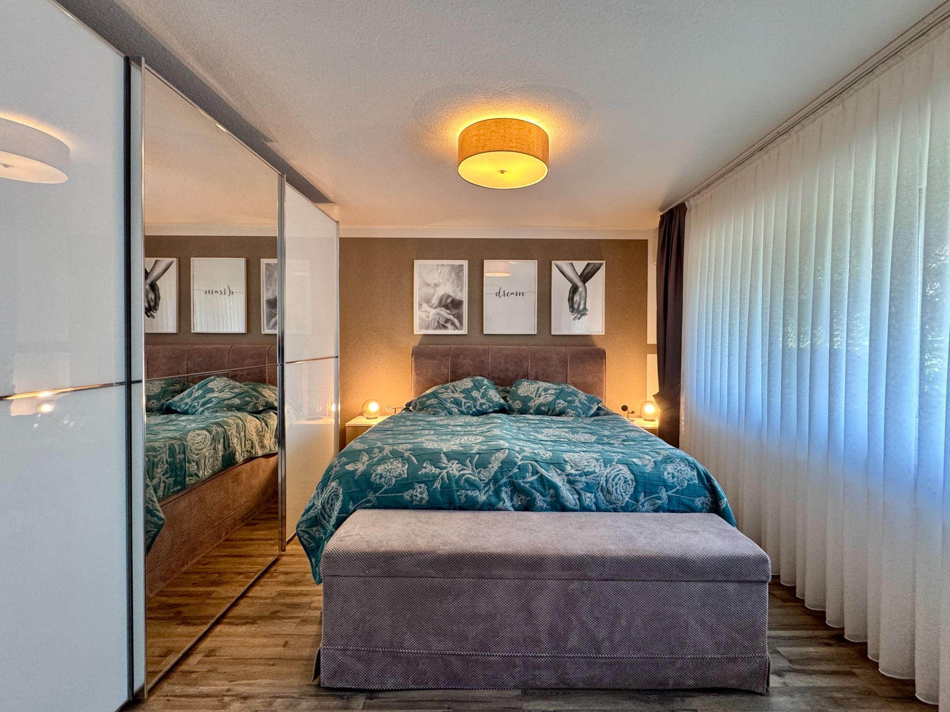 Schlafzimmer mit Doppelbett, großer Spiegel-Schrank und Fenster mit weißen Vorhängen.