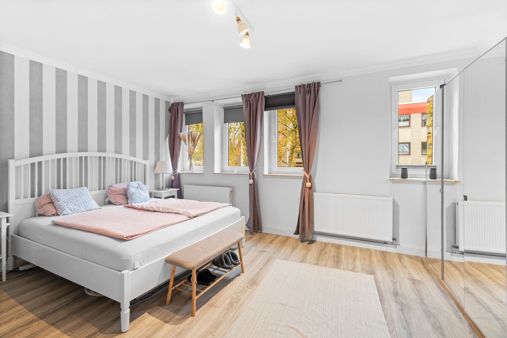 Helles Schlafzimmer mit Doppelbett, Holzbank, Spiegelwand und drei Fenstern mit lila Vorhängen.