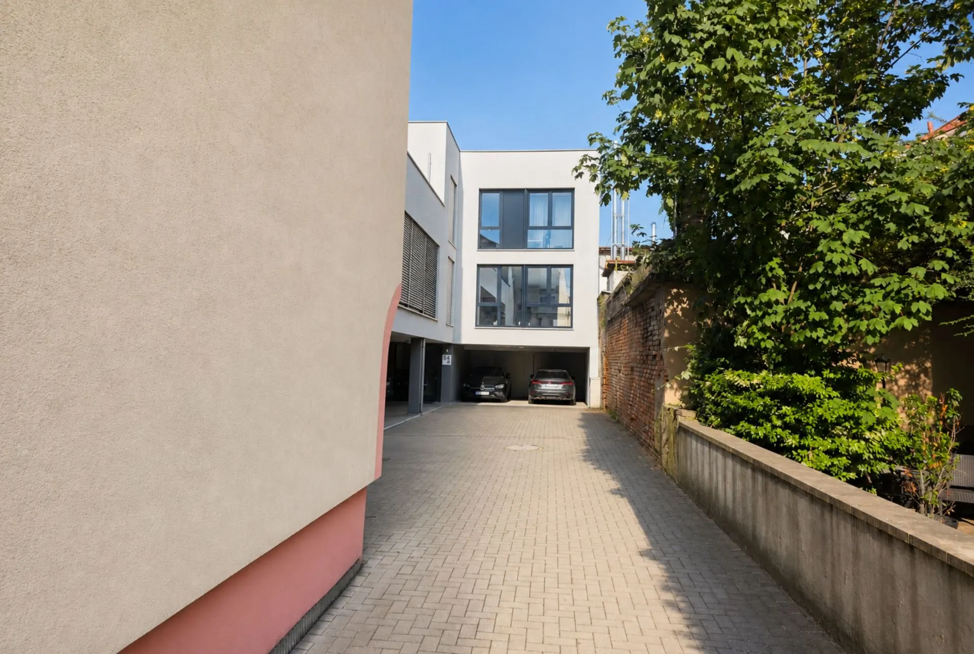 Gepflasterte Einfahrt zu einem modernen Wohngebäude mit zwei geparkten Autos und Baum rechts.