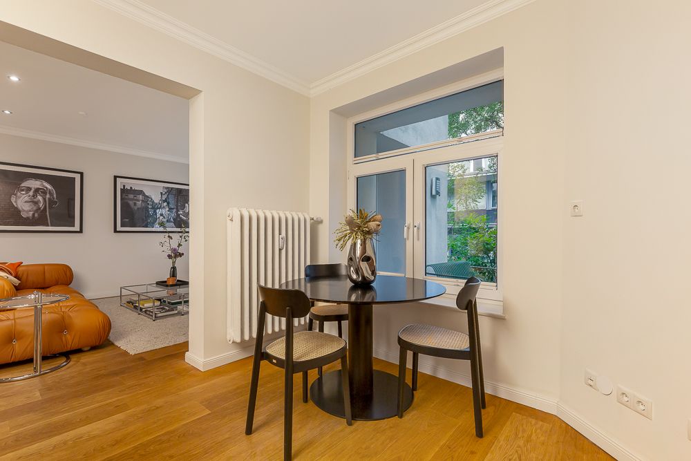 Essbereich mit rundem Tisch, drei Stühlen und Vase am Fenster, Holzboden, Heizkörper an der Wand.