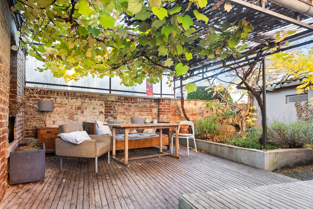 Holzterrasse mit Tisch, Stühlen, Sessel und Pergola mit Weinreben, umgeben von Backsteinmauern.