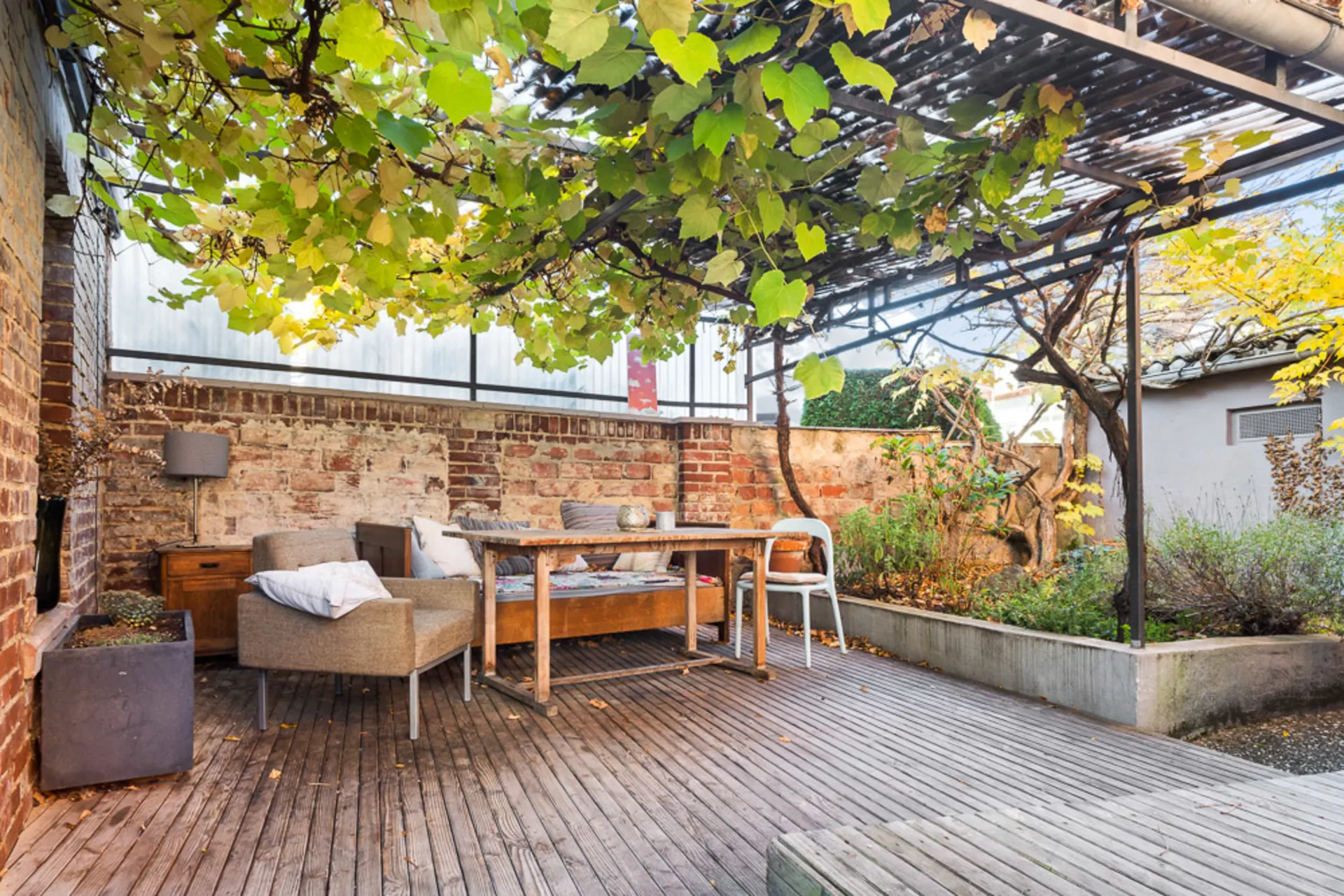 Holzterrasse mit Tisch, Stühlen, Sessel und Pergola mit Weinreben, umgeben von Backsteinmauern.