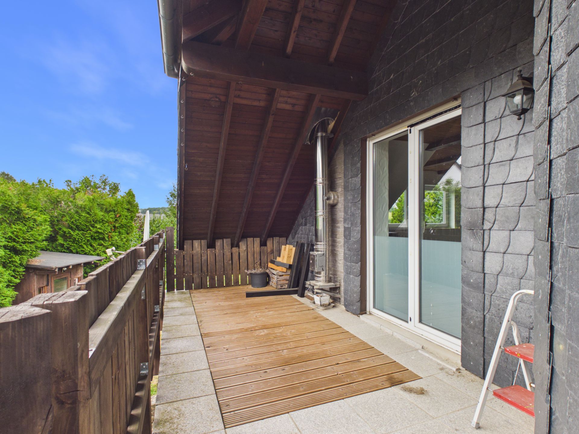 Überdachter Balkon mit Holzgeländer, Holzboden, Holzstuhl und Glasschiebetür.
