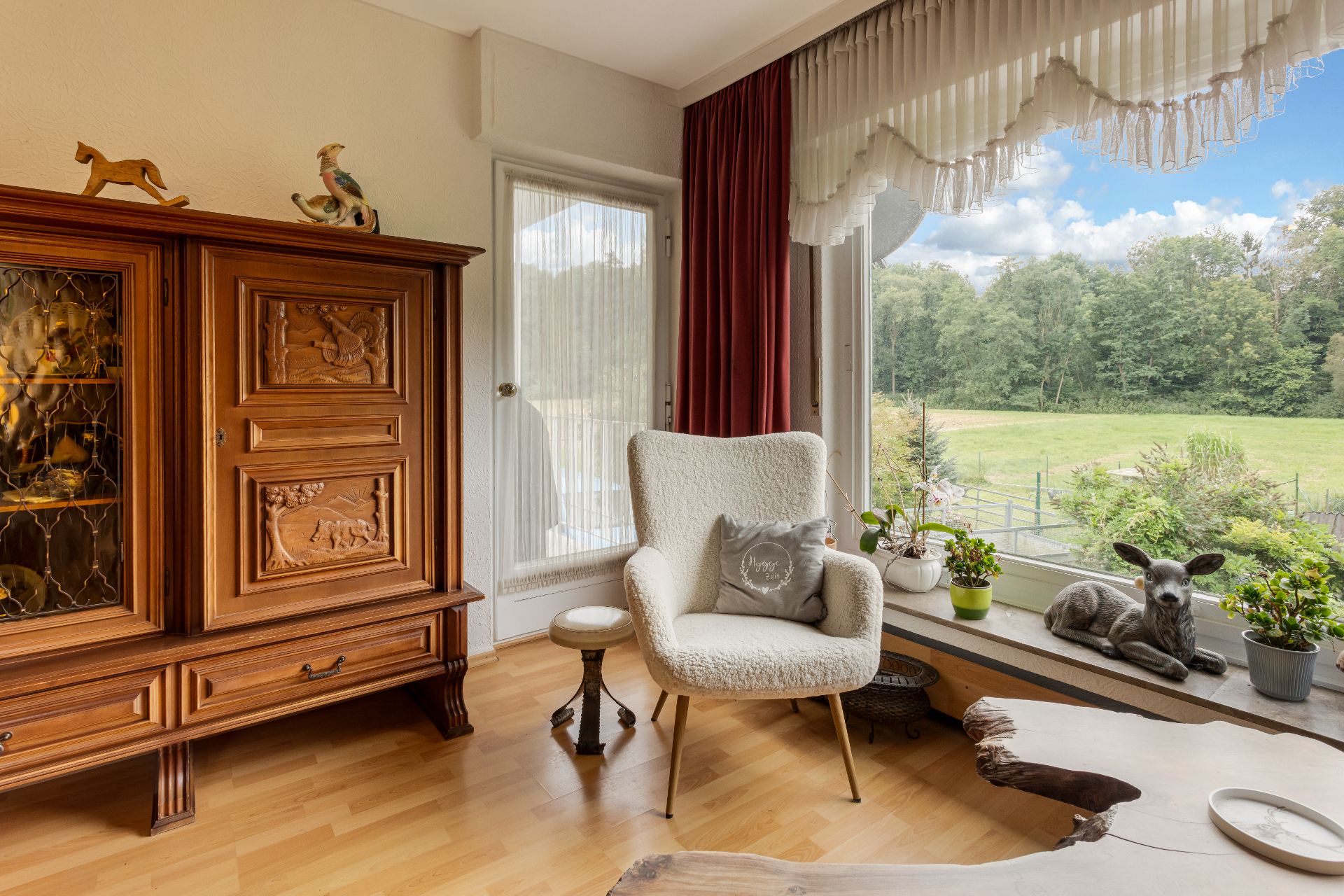 Wohnzimmer mit Holzschrank, Sessel, großem Fenster, Pflanzen und Deko-Reh auf der Fensterbank.
