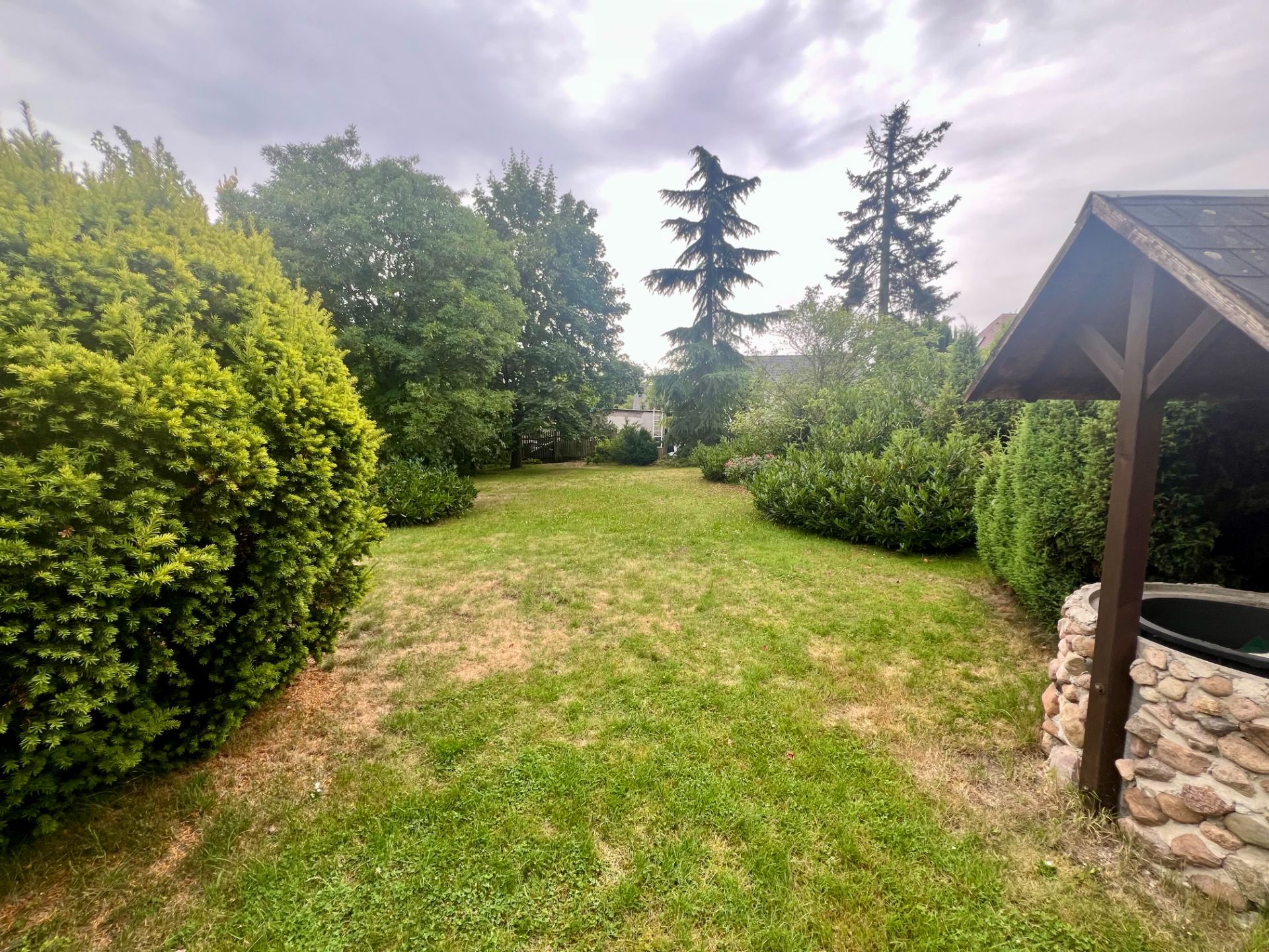 Grüner Garten mit Rasen, Büschen, Bäumen und einem gemauerten Brunnen rechts.
