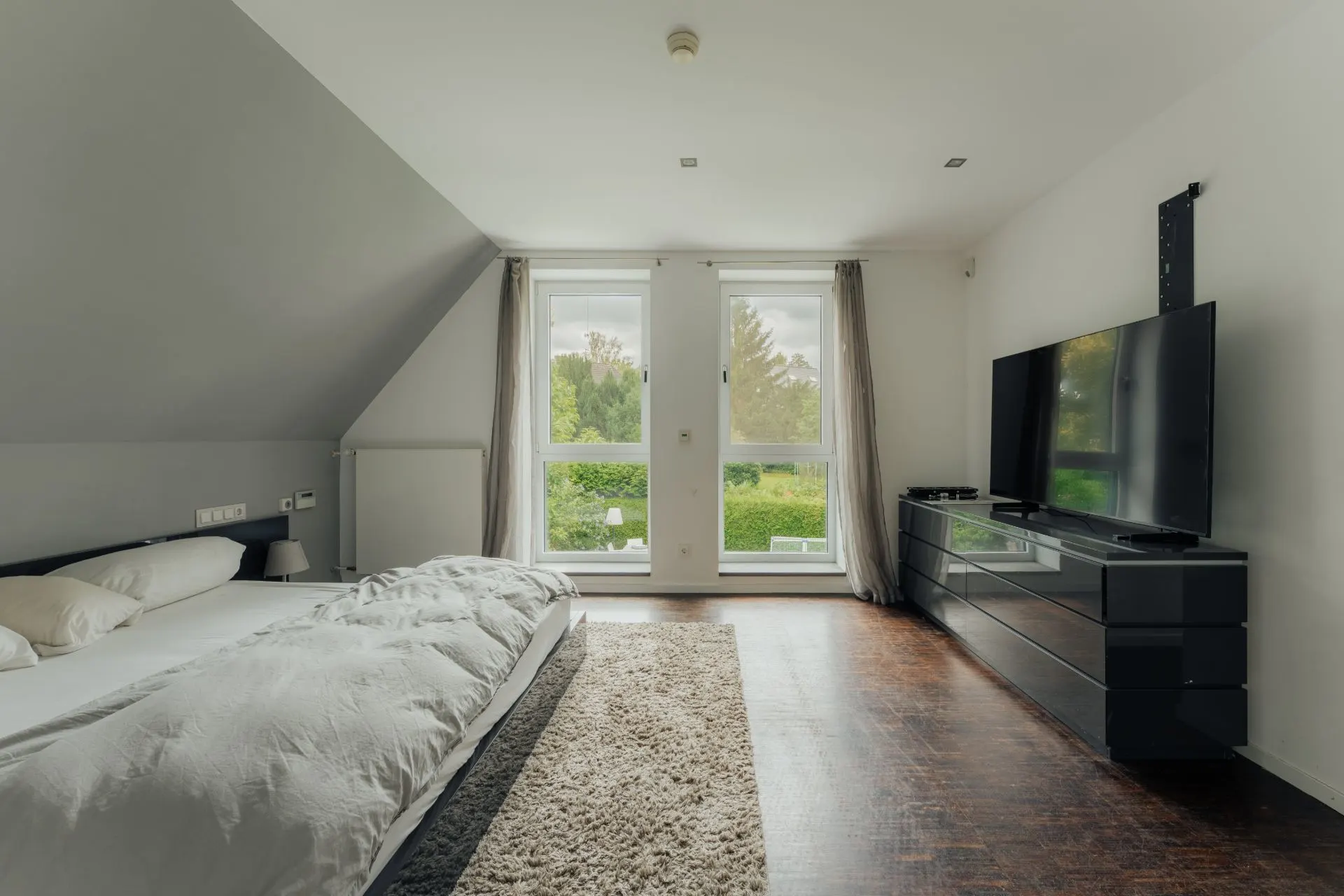 Schlafzimmer mit großem Bett, Teppich, Kommode und Fernseher, zwei Fenster mit Blick ins Grüne.