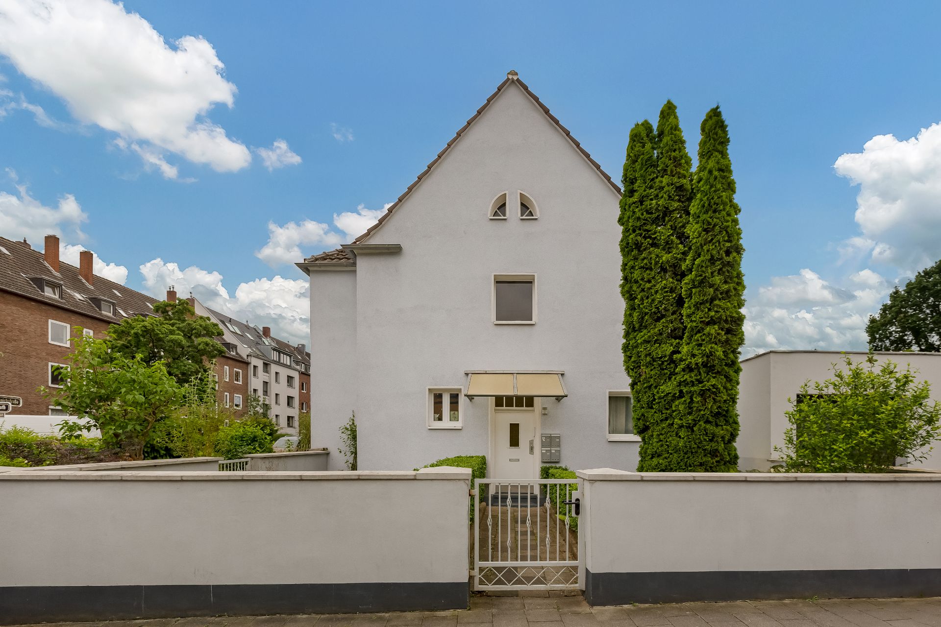Vorderansicht eines weißen Wohnhauses mit Gartenzaun und zwei hohen Bäumen.