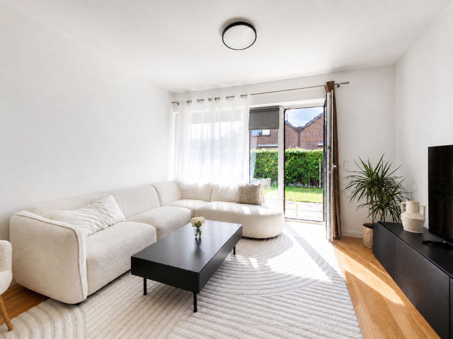 Helles Wohnzimmer mit weißem Sofa, schwarzem Tisch, Teppich und Zugang zum Garten.