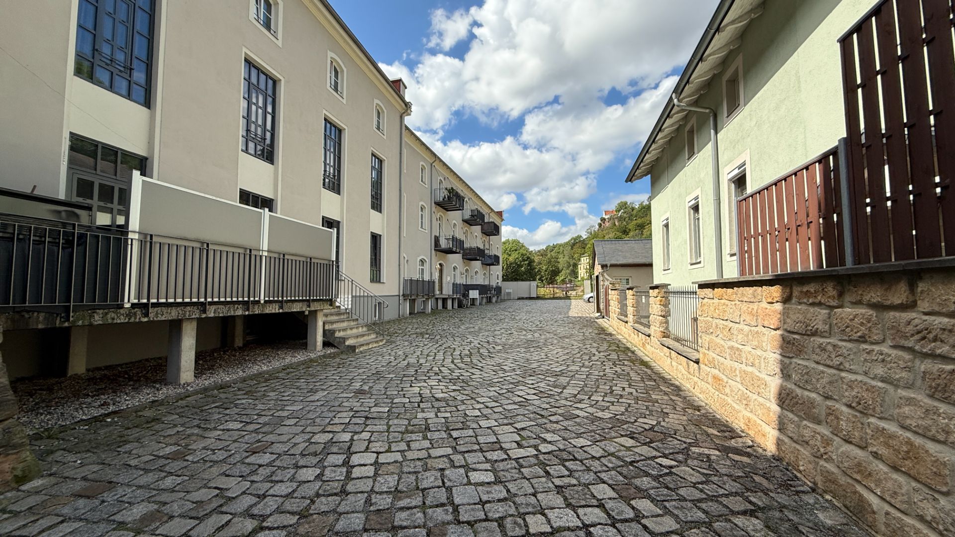 Gepflasterter Weg zwischen zwei Wohngebäuden mit Balkonen und Zäunen, Himmel mit Wolken.