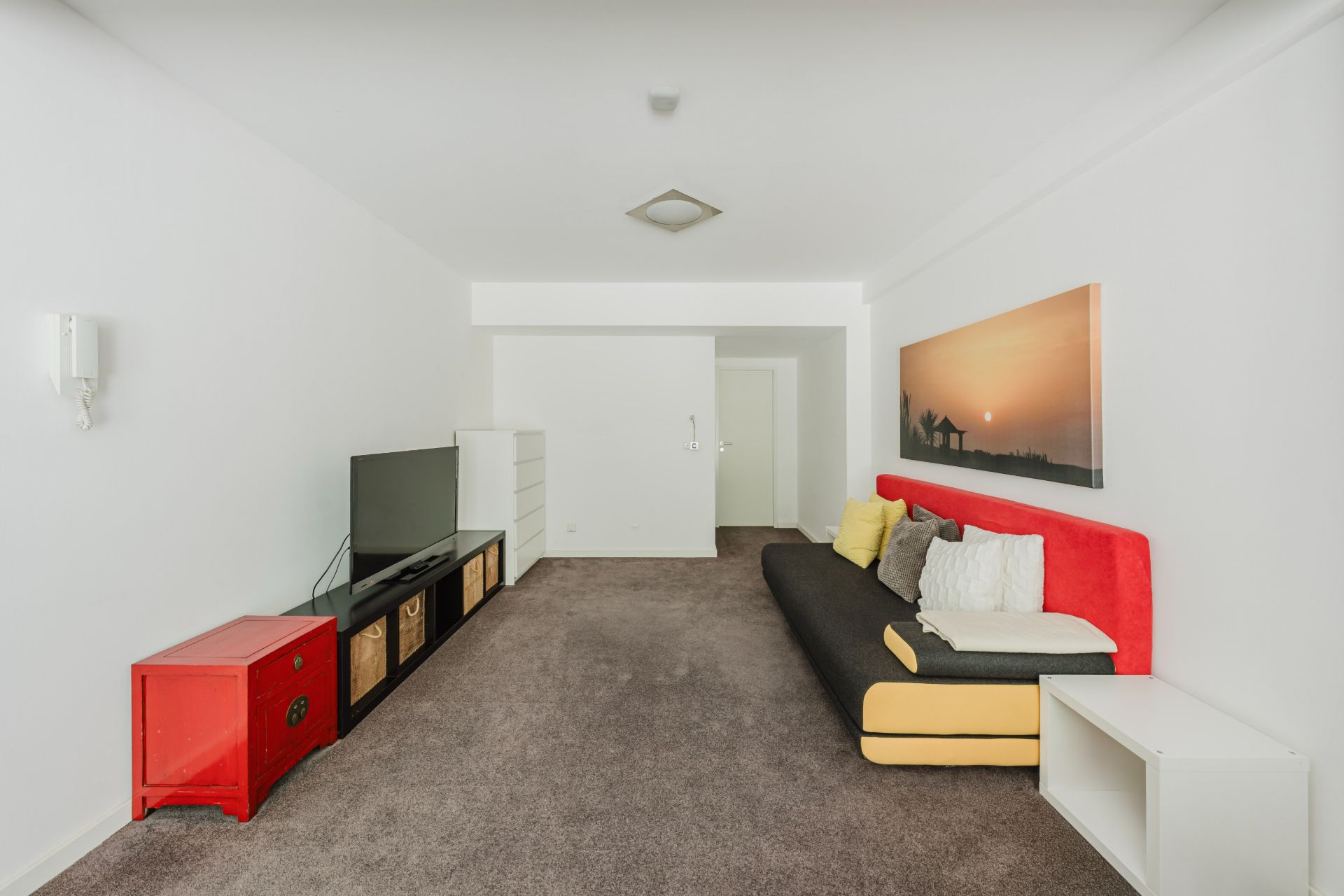 Wohnzimmer mit Sofa, TV, rotem Schrank und Wandbild, Teppichboden und weißen Wänden.