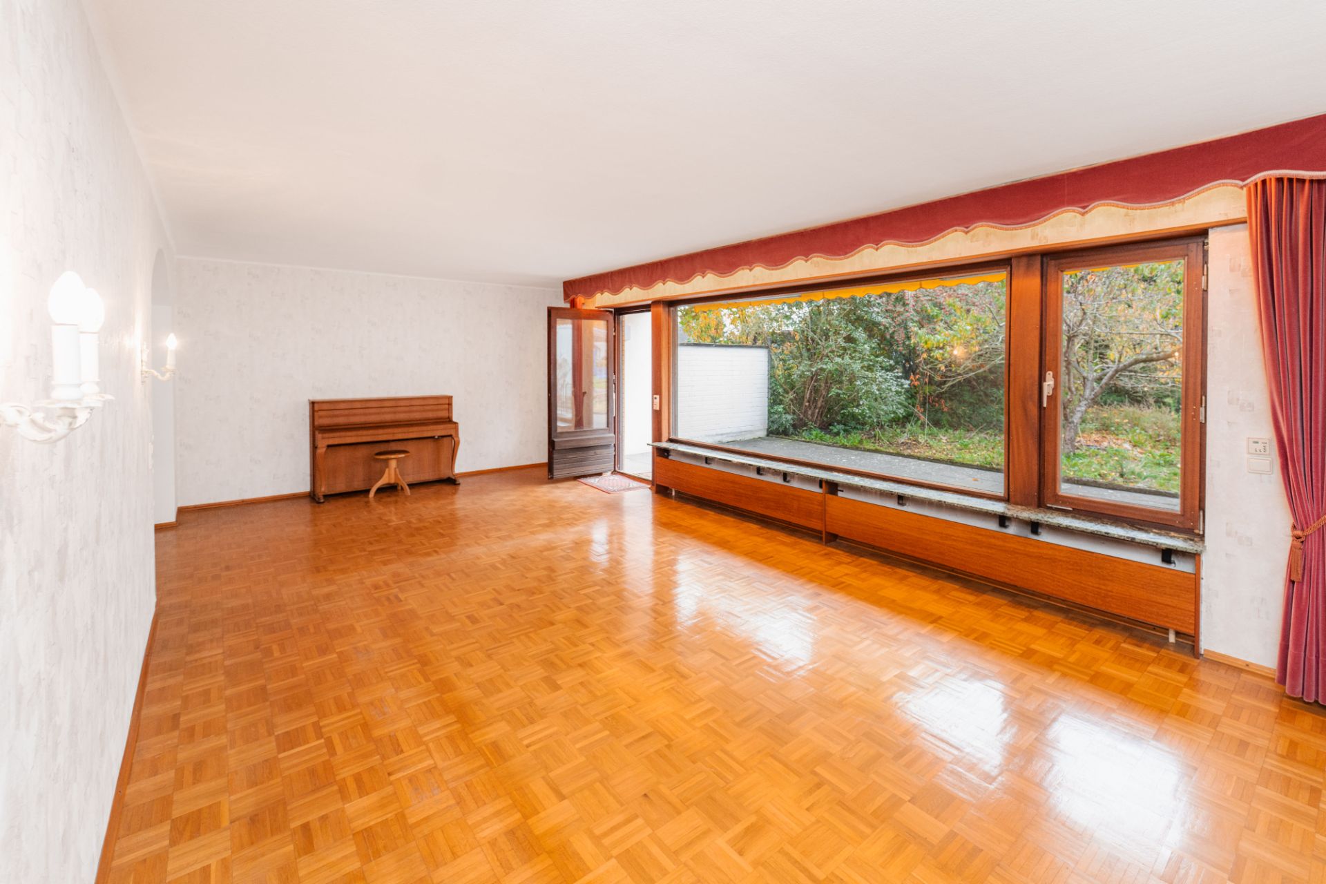 Helles Wohnzimmer mit Parkettboden, großen Fenstern, rotem Vorhang und einem Klavier an der Wand.