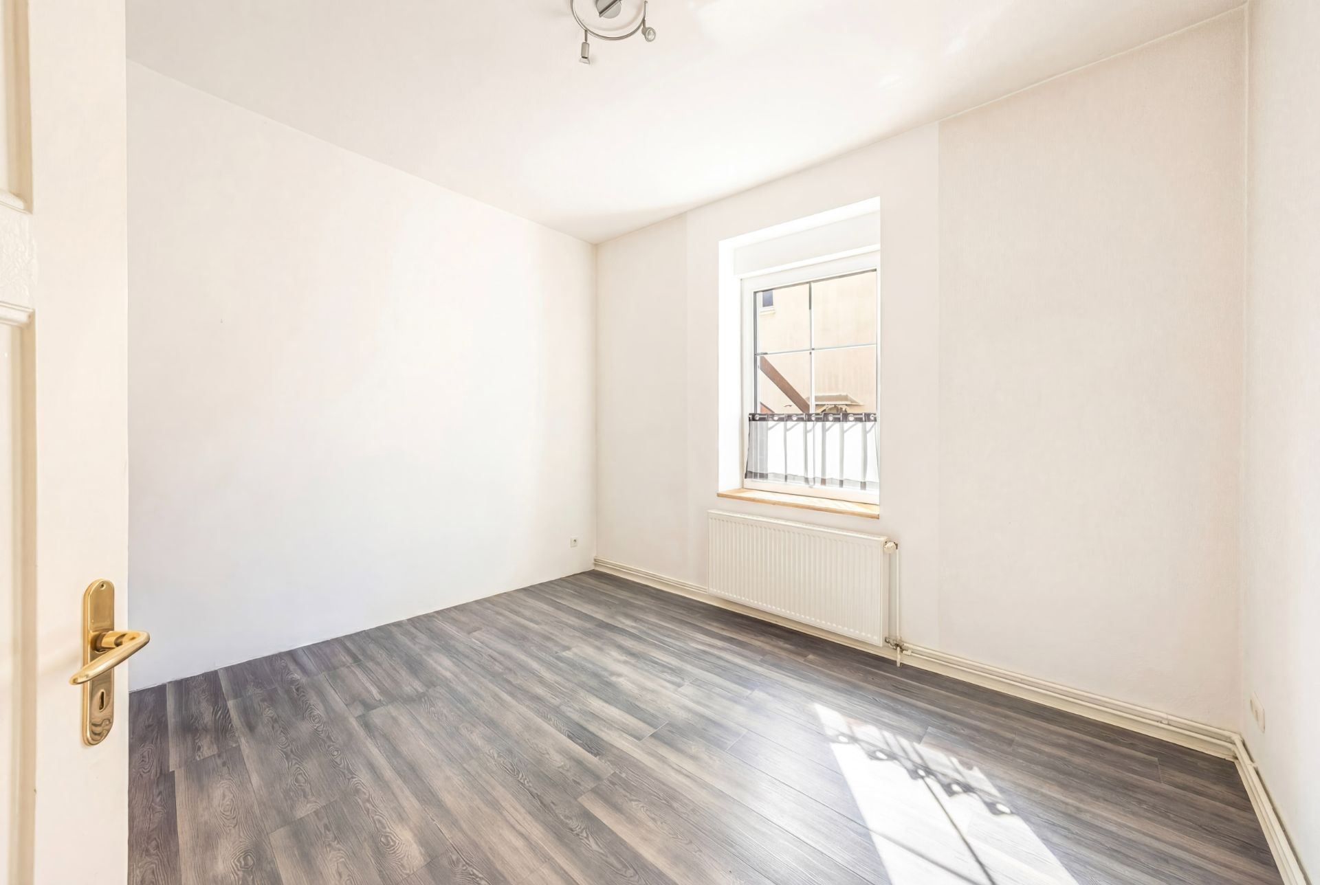 Heller, leerer Raum mit Fenster, hellen Wänden und dunklem Laminatboden.