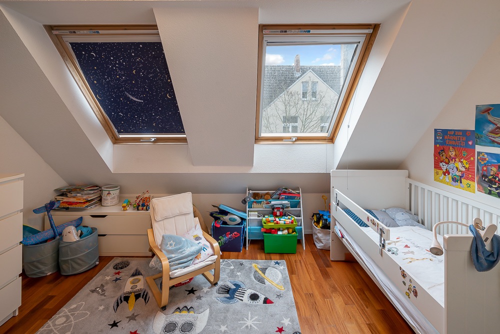 Helles Kinderzimmer mit Dachfenstern, Bett, Spielzeugkisten und Stuhl auf Holzboden.