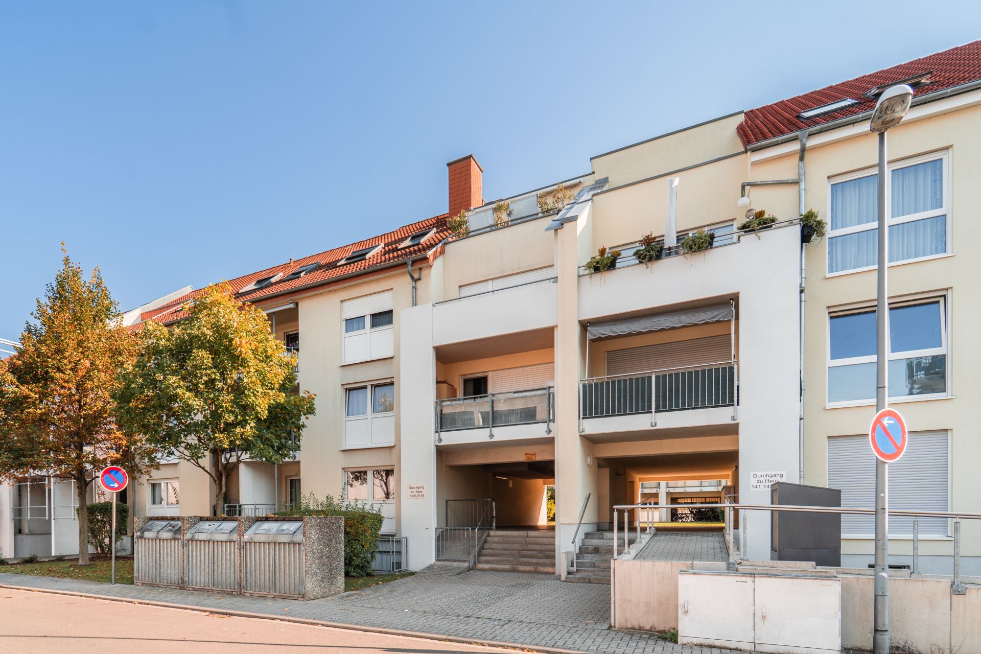 Wohnhaus mit mehreren Balkonen, großen Fenstern und rotem Dach, Bäume und Parkverbotsschilder davor.