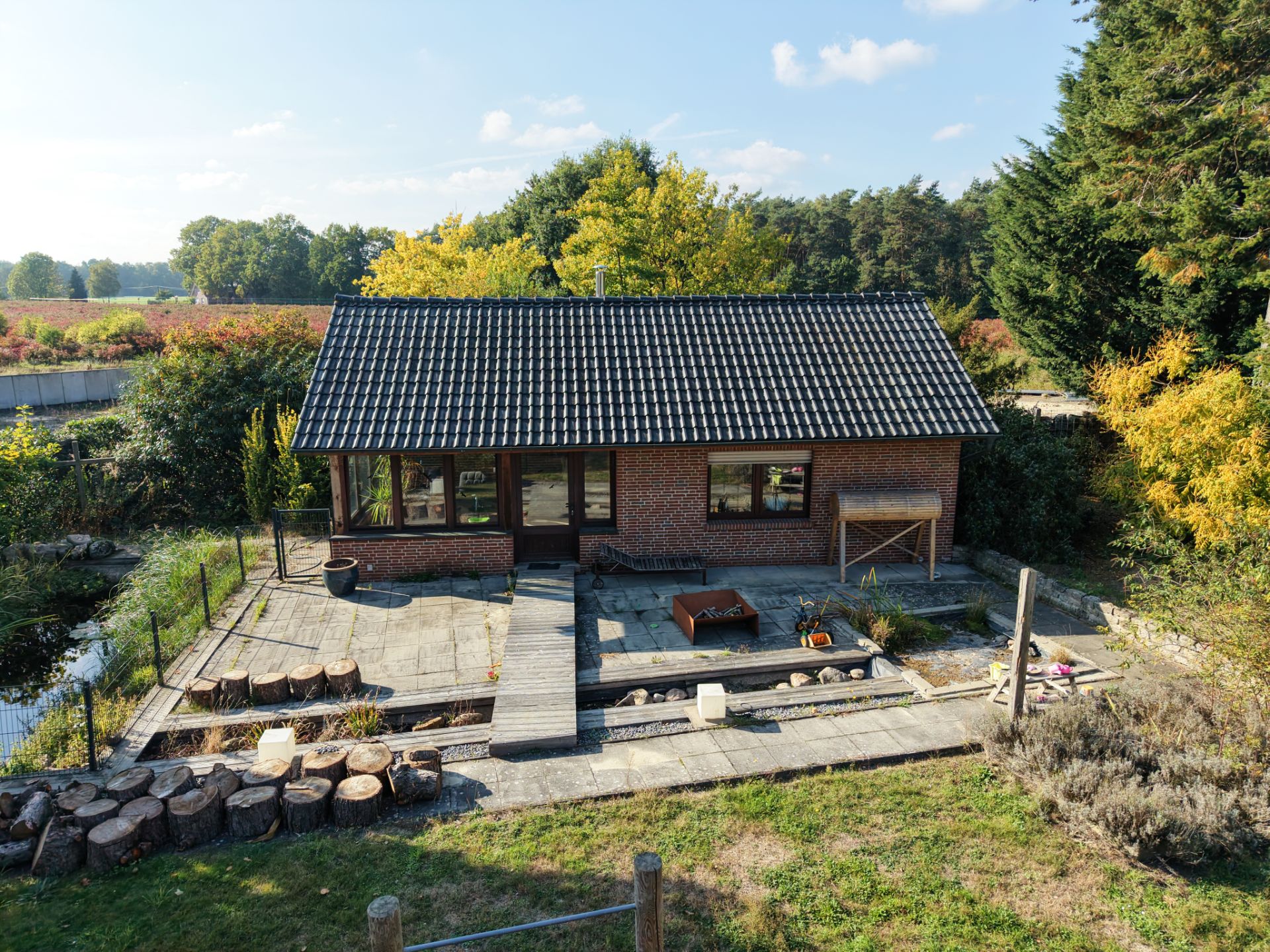 Backsteinhaus mit dunklem Dach und Terrasse im Garten, umgeben von Bäumen und Natur.